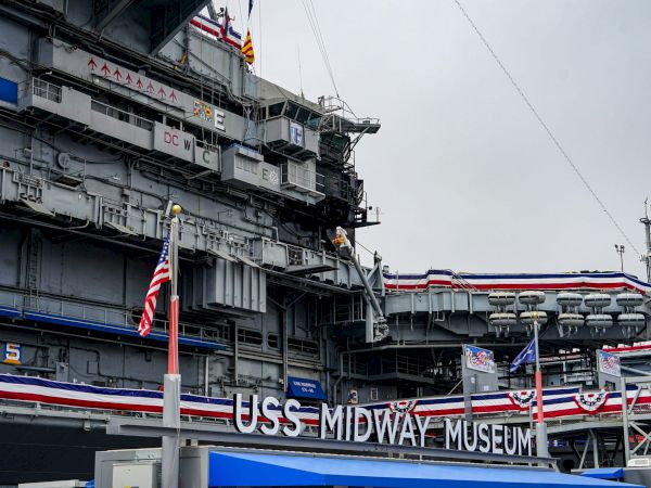USS Midway Museum aircraft carrier with flags, stripes, and visitors aboard, retro navy decor and signage on a gray day.