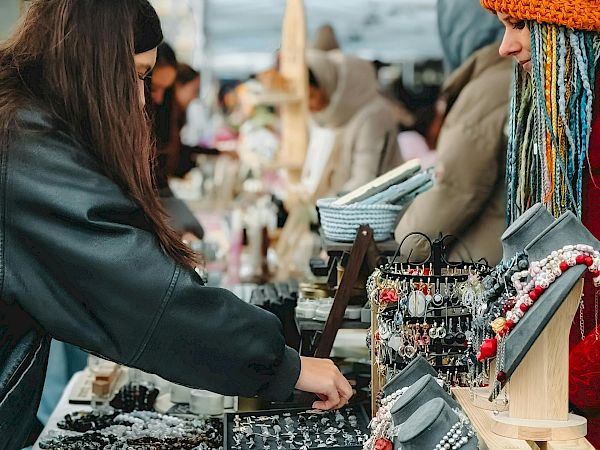 People browsing a street market stall with handmade jewelry and accessories under a canopy; a vendor assists customers, colorful necklaces and bracelets on display.