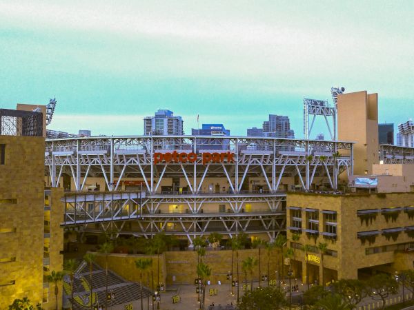 A busy stadium bridge spans between brick and concrete buildings, with tall city skyline in the background under a pale blue sky.