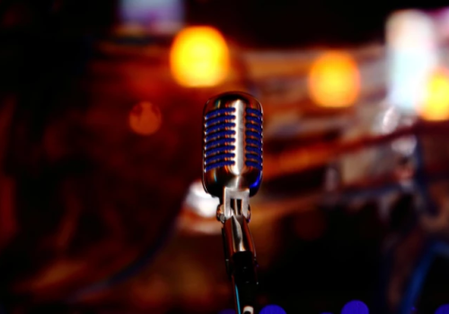 A close-up of a vintage microphone on a stand, spotlighted in a warm, blurred stage setting with colorful bokeh lights in the background.