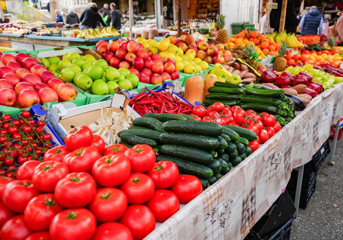 Colorful market stalls with fresh produce: tomatoes, peppers, cucumbers, apples, and other fruits arranged in crates and baskets along a busy outdoor street.