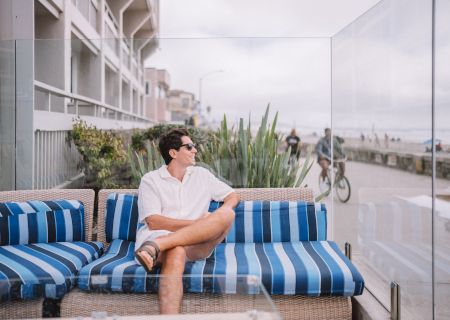 A man in a white shirt sits on a blue striped outdoor sofa on a balcony near the beach, looking away as a sidewalk and cyclists pass by.