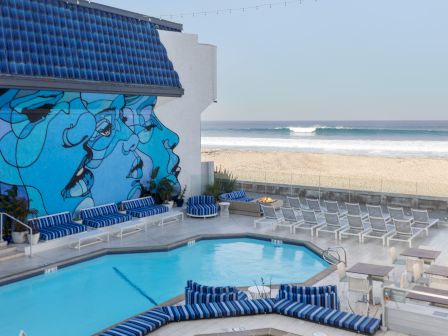 A seaside hotel pool area with blue-and-white lounge chairs, a mural on the wall, and a sandy beach and ocean in the background.