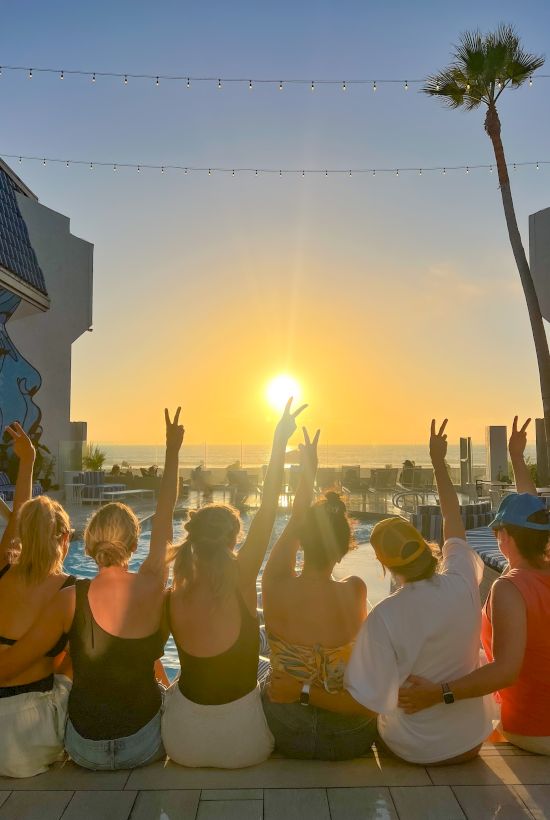 Group of friends at sunset by a pool, arms raised in cheers, ocean in the background, palm trees, vacation vibes.