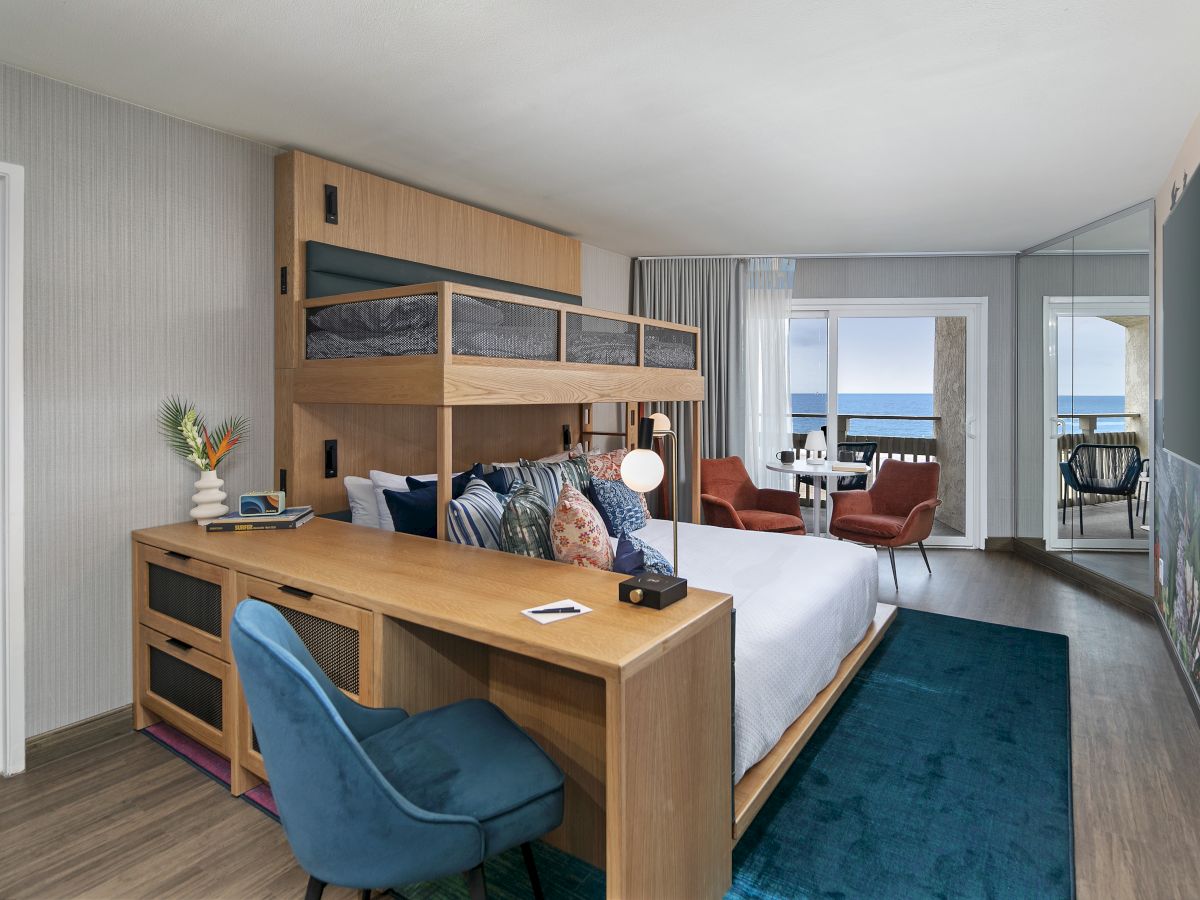 A modern hotel room with a wooden bunk bed, a desk, blue chair, a large wall TV, a teal rug, and a seating area by the window, bright and airy.