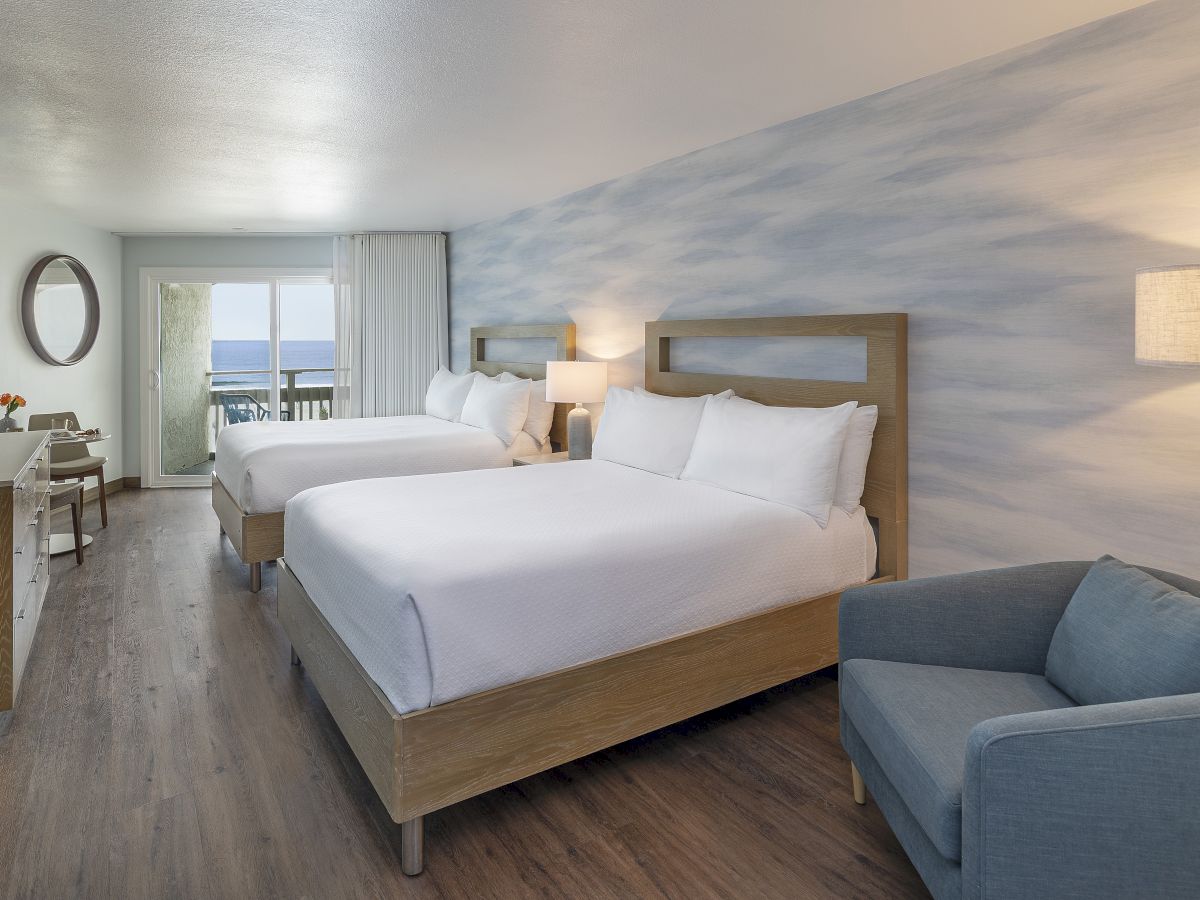 A bright hotel room with two queen beds, a blue accent wall, a seating area, desk, flat-screen TV, and a balcony view.