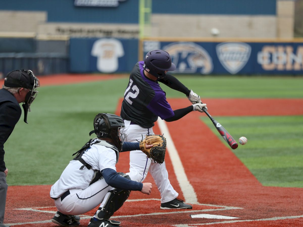 A baseball batter in purple swinging at a pitch, catcher crouched behind home plate, umpire nearby on a red-dirt field.